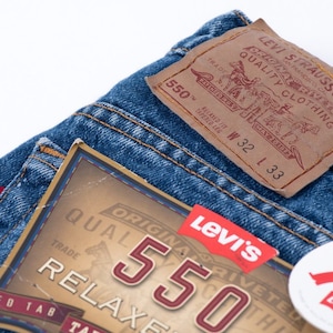 Może przedstawiać: Zbliżenie na parę niebieskich dżins&oacute;w z brązową sk&oacute;rzaną naszywką Levi's z napisem "Original Riveted Quality Clothing" i "550" oraz metką z rozmiarem "W 32 L 33".