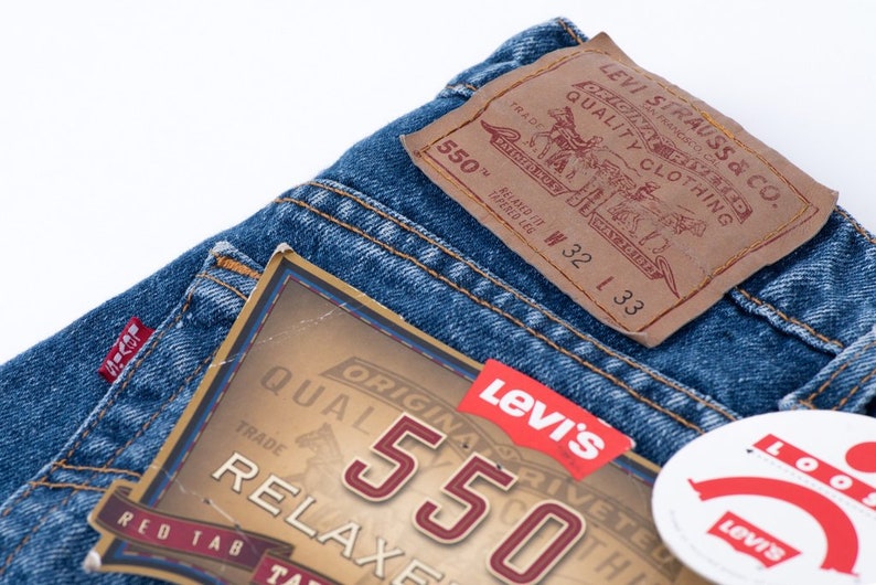 Może przedstawiać: Zbliżenie na parę niebieskich dżins&oacute;w z brązową sk&oacute;rzaną naszywką Levi's z napisem "Original Riveted Quality Clothing" i "550" oraz metką z rozmiarem "W 32 L 33".