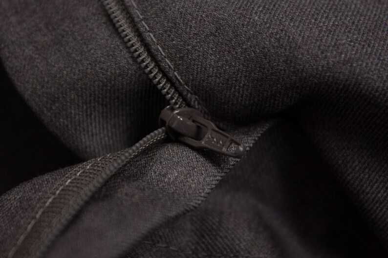 Puede incluir: Primer plano de una cremallera gris oscuro sobre una superficie de tela. La cremallera est&aacute; cerrada y la tela es texturizada.