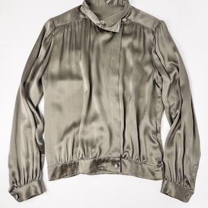 Pode incluir: Uma blusa de seda verde oliva de manga comprida com gola alta e fecho de botão. A blusa tem um caimento solto e uma cintura ligeiramente franzida.