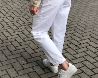 Może przedstawiać: Osoba w białych dżinsach i beżowej kurtce z brązowym swetrem pod spodem. Nosi białe buty sportowe ze wzorem skóry węża.