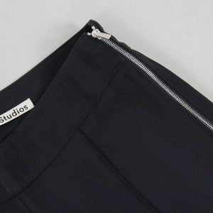 Puede incluir: Primer plano de una secci&oacute;n de tela negra con una cremallera plateada en el borde. La etiqueta "Acne Studios" es visible. La tela es de color oscuro y liso.