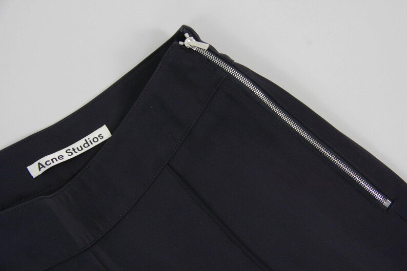 Puede incluir: Primer plano de una secci&oacute;n de tela negra con una cremallera plateada en el borde. La etiqueta "Acne Studios" es visible. La tela es de color oscuro y liso.