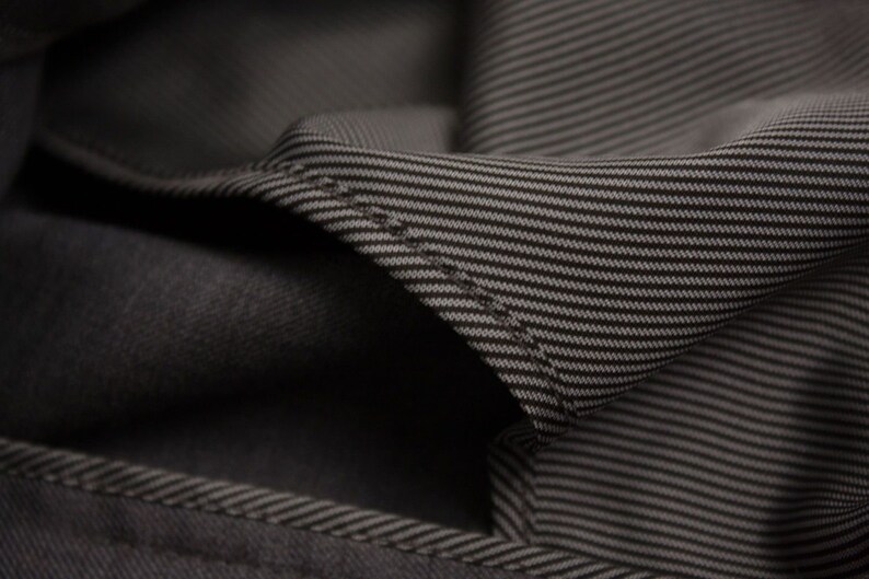 Puede incluir: Primer plano de una tela gris oscura con un sutil patr&oacute;n a rayas. La tela est&aacute; doblada, revelando el reverso del material.