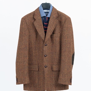Puede incluir: Una blazer de tweed marrón con coderas, combinada con una camisa azul y una corbata a rayas. La blazer tiene un diseño clásico con tres botones y dos bolsillos delanteros, colgada de una percha blanca.