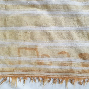 May include: A cream-colored fabric with white stripes and a brown stain that reads "Man". The fabric has a frayed edge and is decorated with small red and brown floral embellishments.