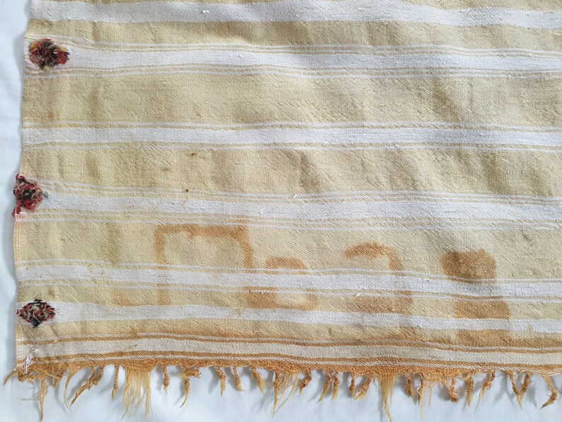 May include: A cream-colored fabric with white stripes and a brown stain that reads "Man". The fabric has a frayed edge and is decorated with small red and brown floral embellishments.