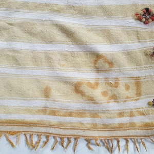 May include: A white and beige striped fabric with a fringe border. The fabric has a brown stain and is decorated with colorful tassels.