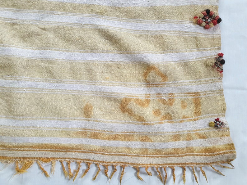 May include: A white and beige striped fabric with a fringe border. The fabric has a brown stain and is decorated with colorful tassels.