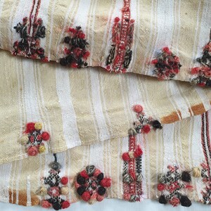 May include: A beige and white striped fabric with red, black, and yellow woven details. The fabric has a fringed edge and is folded in half.