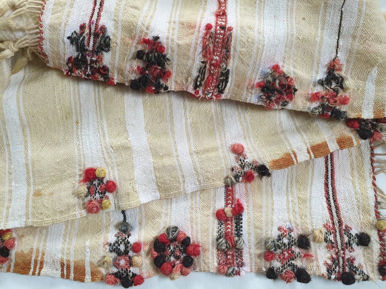 May include: A beige and white striped fabric with red, black, and yellow woven details. The fabric has a fringed edge and is folded in half.
