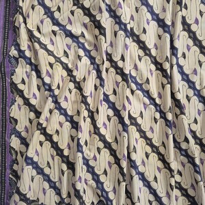 Könnte beinhalten: Ein Textilstoff mit einem sich wiederholenden diagonalen Batikmuster in den Farben Dunkelblau, Creme und Violett. Das Design besteht aus ineinandergreifenden S-förmigen Motiven und kreisförmigen Elementen mit einem vertikalen Randstreifen.