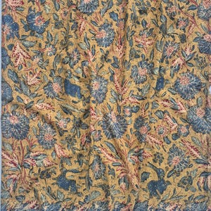 Op de afbeelding: Een stof met een mosterdgele achtergrond en een dicht patroon van blauwe bloemmotieven, groene bladeren en kleine rode accenten. Het ontwerp bevat gestileerde blauwe konijnen die verspreid tussen de bloemen en het gebladerte staan.