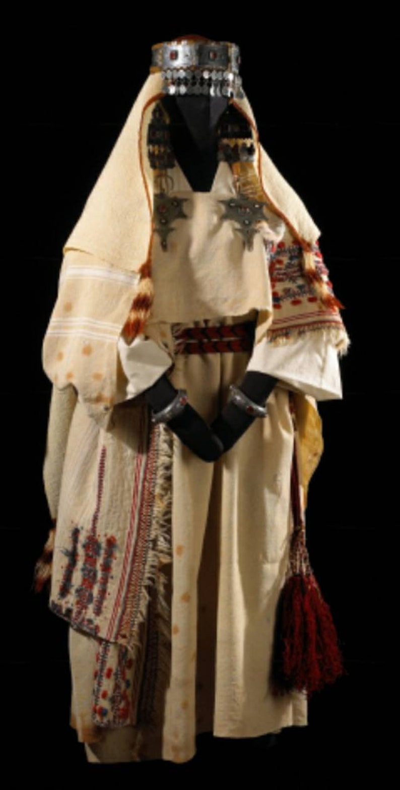 May include: A white robe with red and blue embroidery, a silver headpiece with red jewels, and a red tassel. The robe is worn over a white tunic with silver embellishments.