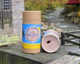 Op de afbeelding: Een bruine kartonnen buis met een blauw etiket waarop staat "BARE ELEMENTS Vegan Sun Stick be better, be bare With Non-Nano Zinc Oxide". De buis ligt naast een bruine buis met een gat in de bovenkant en een lichtbruine substantie erin.