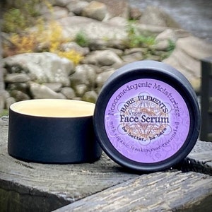 May include: A small black jar with a purple label that reads "Bare Elements Vegan Face Serum with organic frankincense essential oil. Be better, be bare." The jar is sitting on a wooden surface.