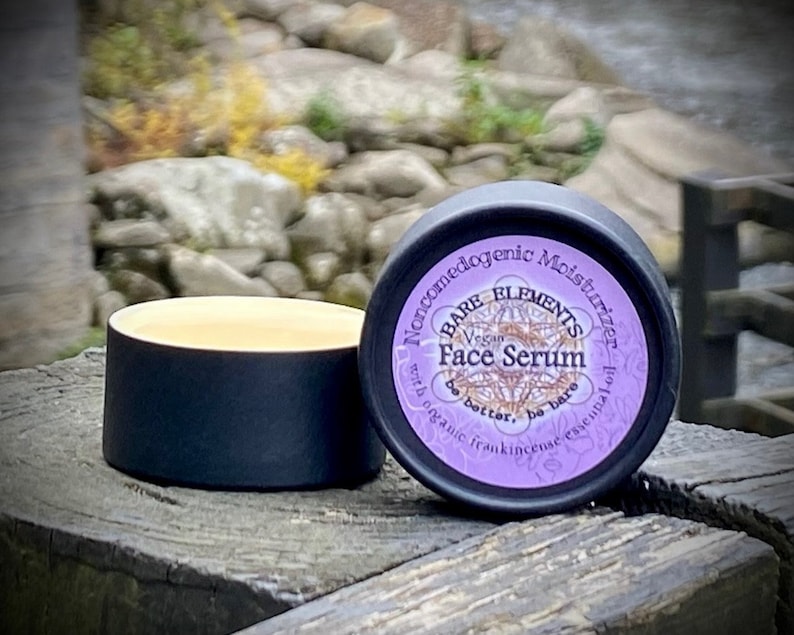 May include: A small black jar with a purple label that reads "Bare Elements Vegan Face Serum with organic frankincense essential oil. Be better, be bare." The jar is sitting on a wooden surface.