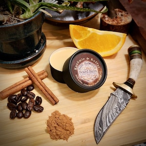 Op de afbeelding: Een klein zwart potje met een wit etiket waarop staat "Kashmir & Koffee BARE ELEMENTS Beard Balm & After Shave Lotion with coffee, cinnamon, more..." Het potje staat op een houten oppervlak met koffiebonen, kaneelstokjes en een klein hoopje kaneelpoeder.