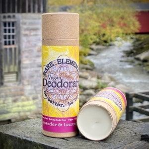 May include: A brown cardboard tube with a yellow and pink label that reads "Bare Elements Vegan Deodorant be better, be bare" and "Lavender & Lemon". The tube is sitting on a wooden surface with a stream of water in the background.