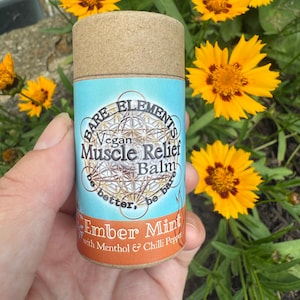 May include: A cylindrical container of Bare Elements Vegan Muscle Relief Balm, featuring a light blue label with gold text and a brown cap. The label reads "Ember Mint" with menthol and chili pepper. Yellow flowers with brown centers are in the background.