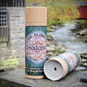 May include: Two cardboard tubes of vegan deodorant, one with the label "BARE ELEMENT Vegan Deodorant be better, be ba Tea Tree & Rose" visible. The tubes are on a wooden surface with a blurred background of a stream and a red bridge.