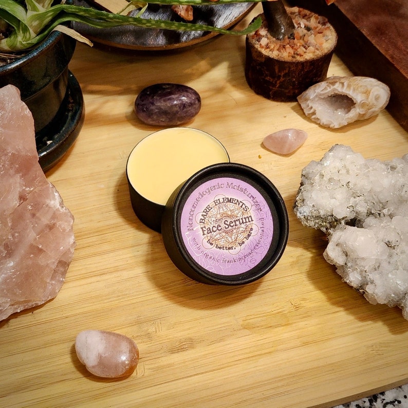 May include: A small, round jar of face serum with a purple label that reads "Bare Elements Face Serum with organic frankincense & myrrh.  The jar is sitting on a wooden surface with several pink and white crystals.