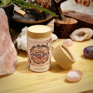 May include: A small, brown cardboard tube with a white label that reads "Bare Elements Vegan Lotion Stick Be better, be bare. Unscented". The tube is sitting on a wooden surface with pink and purple crystals.