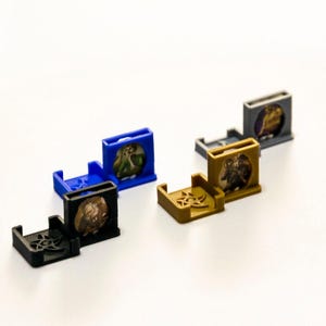 May include: Six small, colourful plastic stands with different designs. Each stand has a small circular space to hold a miniature figure or game piece. The stands are blue, black, grey, and gold.
