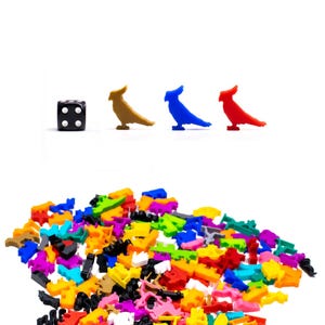 Peut inclure: Un dé noir à six faces avec des points blancs, ainsi que trois figurines d'oiseaux en plastique de couleurs vives. Les oiseaux sont brun, bleu et rouge. Un grand tas de figurines d'oiseaux en plastique de couleurs vives dans diverses couleurs et formes sont éparpillés au bas de l'image.