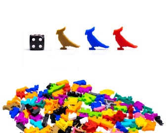 Può includere: Un dado nero a sei facce con punti bianchi, insieme a tre figurine di uccelli in plastica colorate. Gli uccelli sono marroni, blu e rossi. Un grande mucchio di figurine di uccelli in plastica colorate in vari colori e forme sono sparse nella parte inferiore dell'immagine.