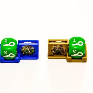 May include: Two green dice with magnifying glass symbols and the numbers 1 and 2 are in holders. One holder is blue and the other is gold.