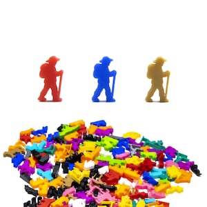 Puede incluir: Tres figuritas de excursionistas en miniatura, una roja, una azul y una marrón, están alineadas en una fila sobre un fondo blanco. Un montón de piezas de plástico de colores están esparcidas en primer plano.