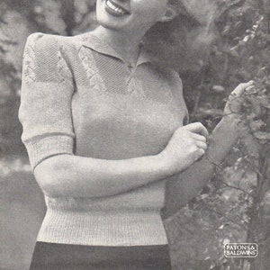 May include: A woman wearing a light grey knitted sweater with a collar and a lace pattern on the sleeves. She is smiling and her hair is blowing in the wind.