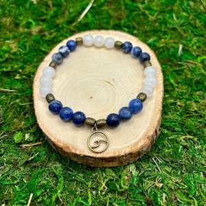 May include: A blue and white beaded bracelet with a bronze wave charm. The bracelet is made with blue sodalite and white howlite beads.