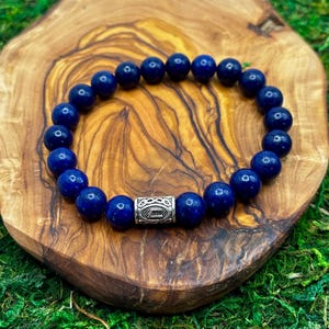 Puede incluir: Una pulsera de cuentas azul oscuro con una cuenta de acento plateada. La pulsera está hecha de cuentas redondas y pulidas. La pulsera se exhibe sobre una superficie de madera con un fondo verde musgo.