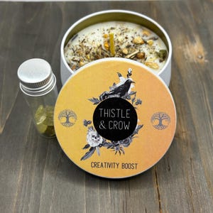 Puede incluir: Una lata de vela blanca con una tapa amarilla que presenta un cuervo negro y un diseño floral. La tapa tiene el texto "Thistle & Crow" y "Creativity Boost". Se incluye un pequeño frasco de vidrio con cristales amarillos.