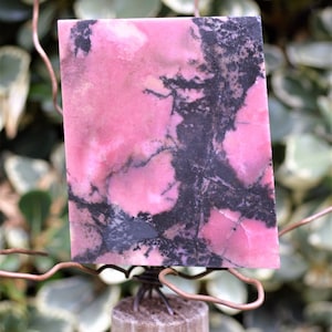 May include: A rectangular slab of pink rhodonite stone with black veining. The polished stone is mounted on a wooden base with copper wire. The background features green foliage.