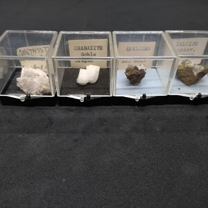 May include: Four clear plastic display cases, each containing a different mineral specimen. The specimens include amethyst, chabazite, analcime, and calcite. Each case has a black base and a small label with the mineral's name.