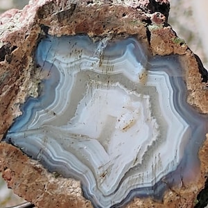 May include: A cross-section of a geode featuring a rough, textured, reddish-brown outer crust surrounding a smooth, translucent interior. The center displays concentric bands of white, light gray, and pale blue mineral deposits in a star-like pattern.