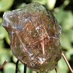Puede incluir: Un gran cristal de cuarzo transparente con inclusiones de cobre. El cristal está en un soporte de metal y tiene una textura rugosa y natural.