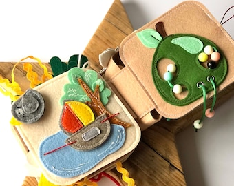 May include: A felt activity book for toddlers with a green apple, a boat on a lake, and a bird. The book has pages with different textures and activities for little hands to explore.