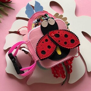 May include: A pink felt book with a ladybug on the front. The ladybug has red wings with black spots and a black body with white eyes. The book has a loop for hanging.