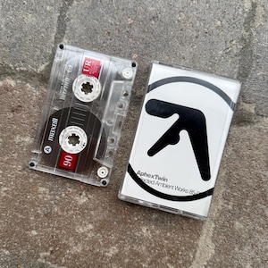 May include: A clear cassette tape with a black and red label, next to a white cassette case with a black logo and the text "Aphex Twin Selected Ambient Works 85-92". The tape is labeled "maxell 90".