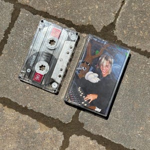 May include: Two clear cassette tapes, one with a Maxell 90 label and the other with a photo of a person wearing headphones and a white shirt, using a mixing board.