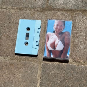 May include: A light blue cassette tape and a clear plastic cassette case featuring a photographic insert of a person with a shaved head wearing a white bikini top against a blue background. The cassette shell is solid light blue plastic.
