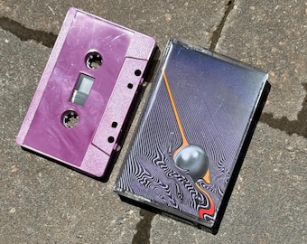 May include: A purple cassette tape case with a black cassette tape inside. The cassette tape has a black and white abstract design with a black sphere in the centre.