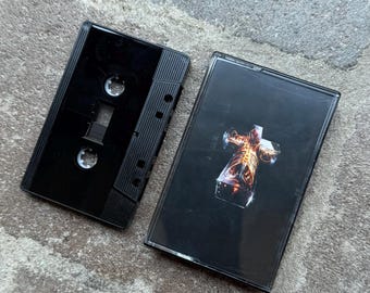 May include: A black cassette tape with a clear case, featuring a dark cover with a stylized, glowing figure. The cassette is open, revealing the tape and its components. The image is set against a gray, textured surface.