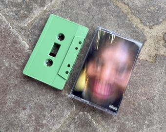 May include: A green cassette tape case with a clear plastic case containing a cassette tape with a blurred image of a person's face. The cassette tape has a Parental Advisory sticker on it.