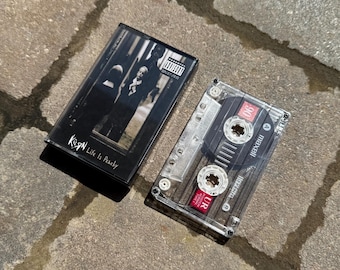 May include: A transparent cassette tape case featuring the album artwork for KoЯn's "Life is Peachy". The cassette tape is visible within the case. The case is open and resting on a stone surface.
