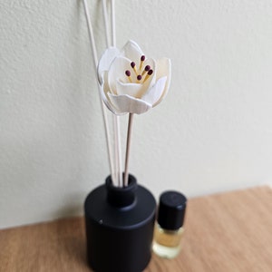 May include: A black diffuser bottle with wooden sticks and a white wooden flower on top. The diffuser bottle is sitting on a wooden surface.
